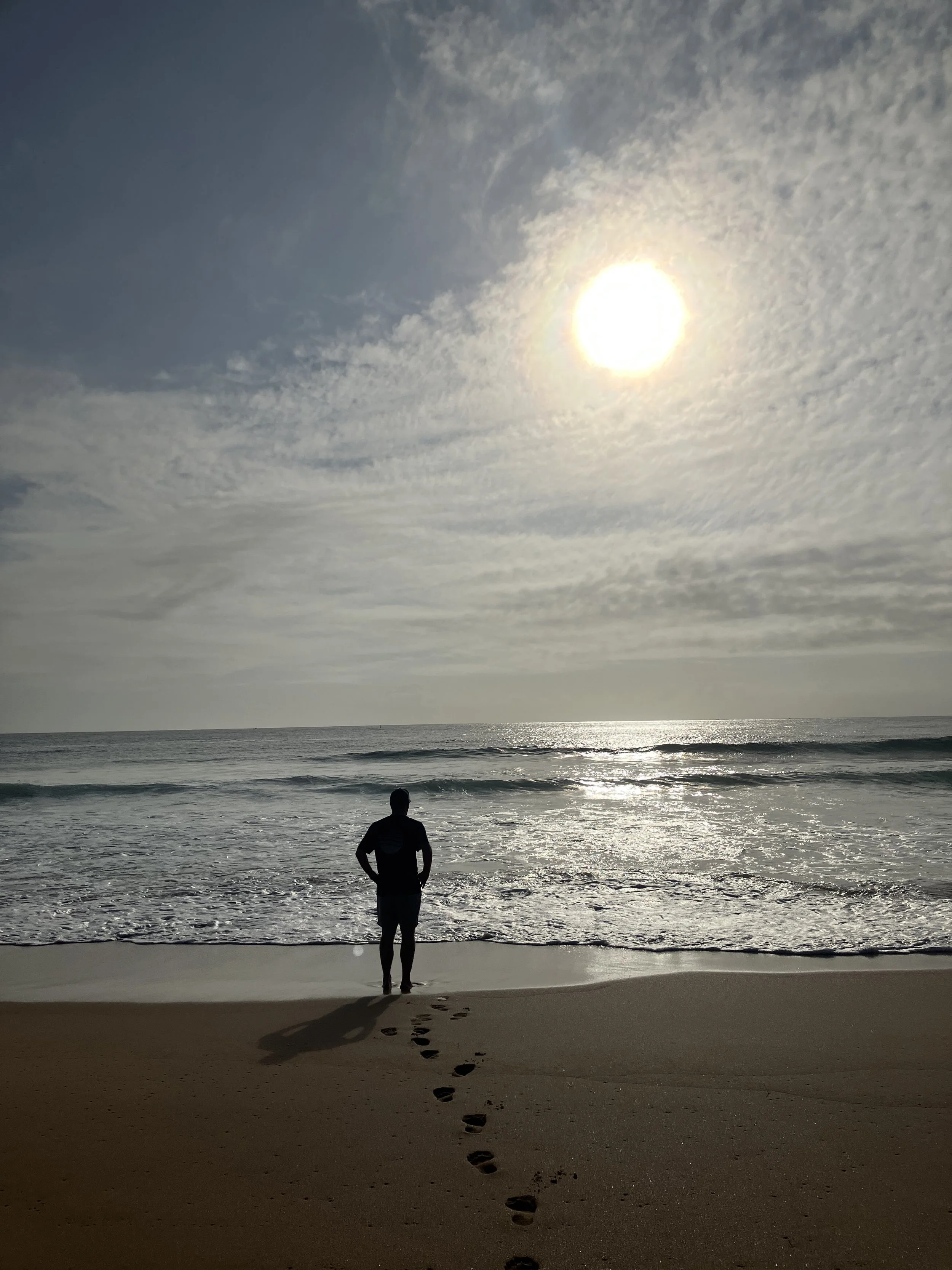 A person standing on a sandy beach, facing the ocean under a bright, partly cloudy sky with the sun overhead. Footprints lead to the person.
