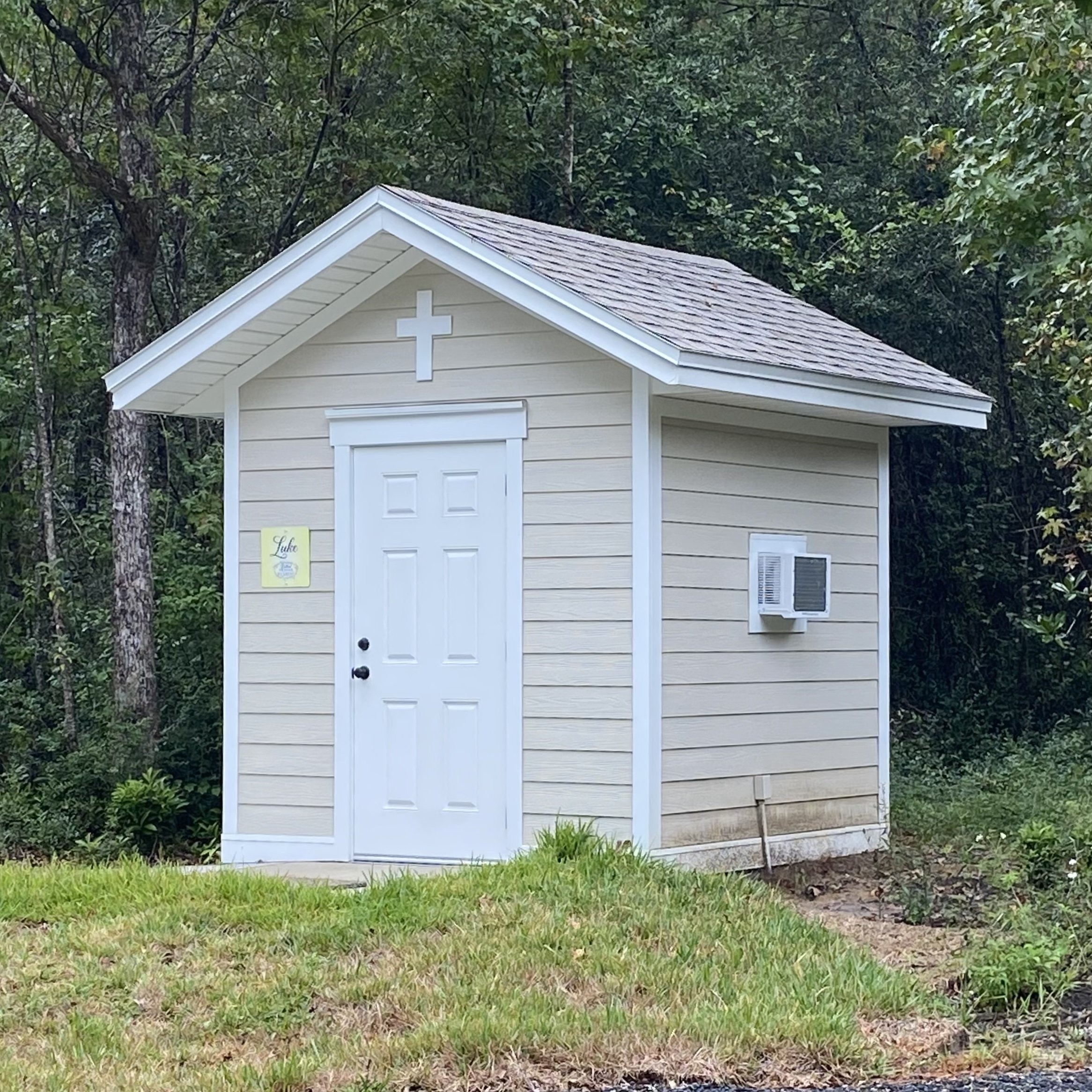 Small beige building with white trim and a cross on the roof, set in a wooded area.
