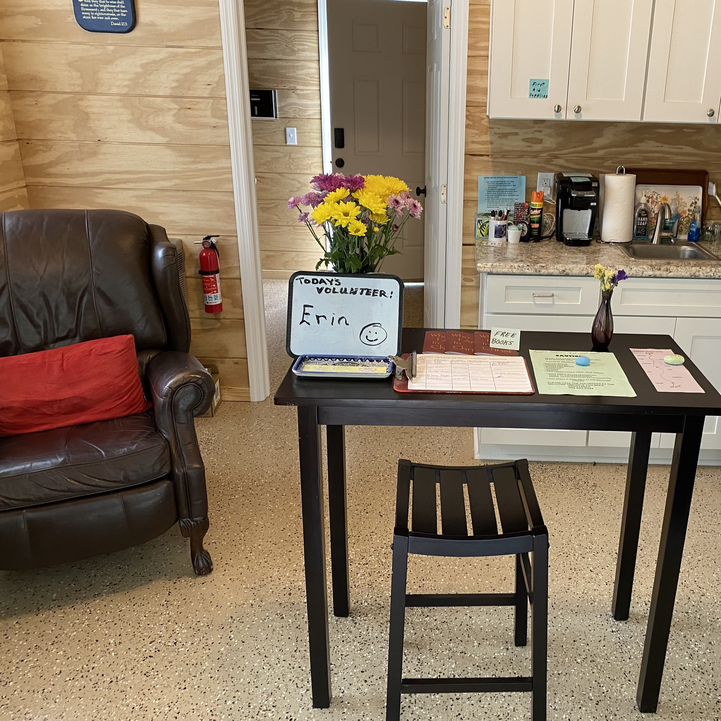 Reception area with a brown leather armchair, red pillow, and a black high table. A whiteboard on the table says "Today's Volunteer: Erin" with a smiley face. The table has flowers, a clipboard, and flyers. A small kitchen area is in the background w