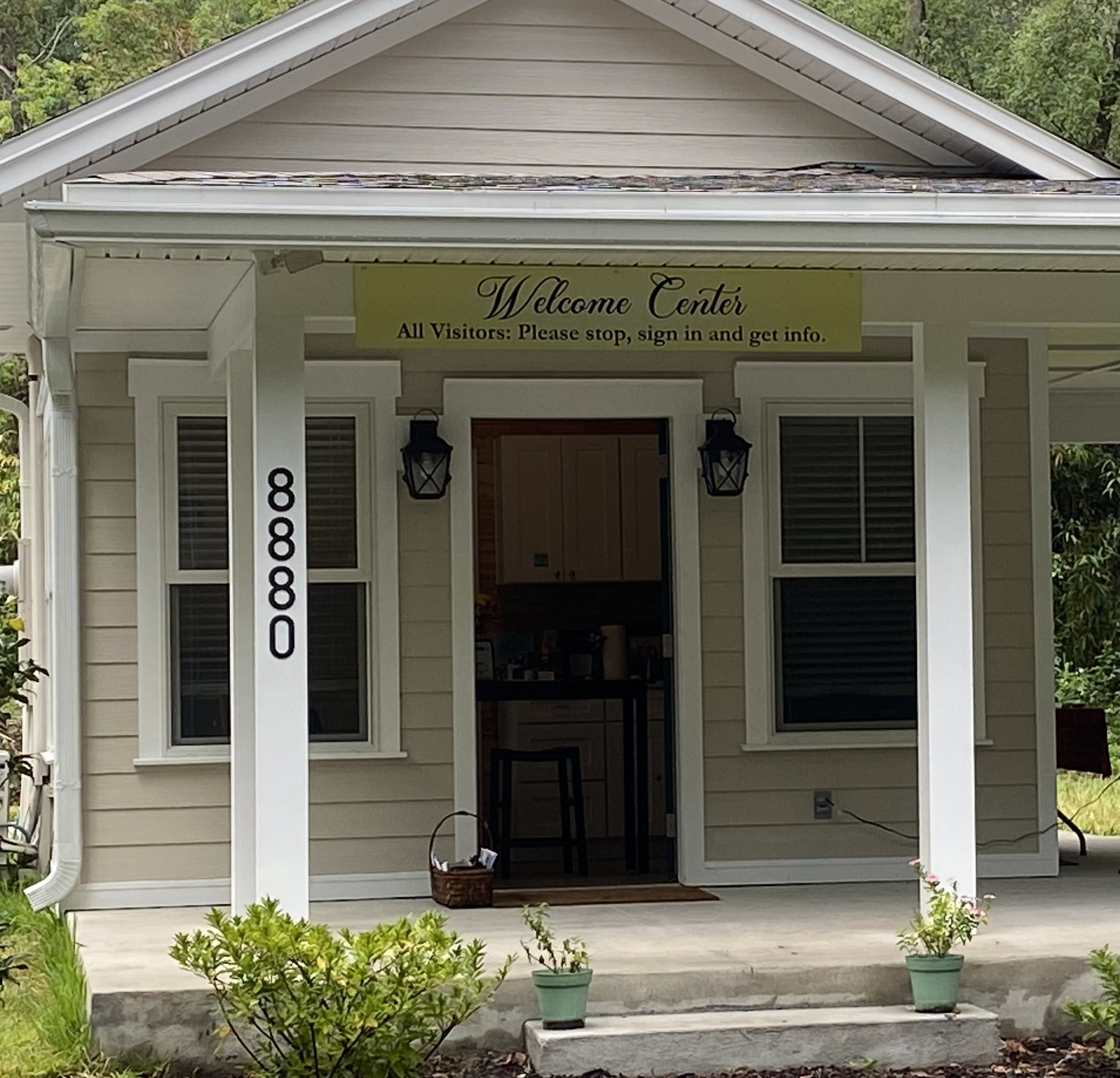 Small building with a sign reading 'Welcome Center' on the porch, address number 8880 displayed on a pillar, surrounded by plants and trees.