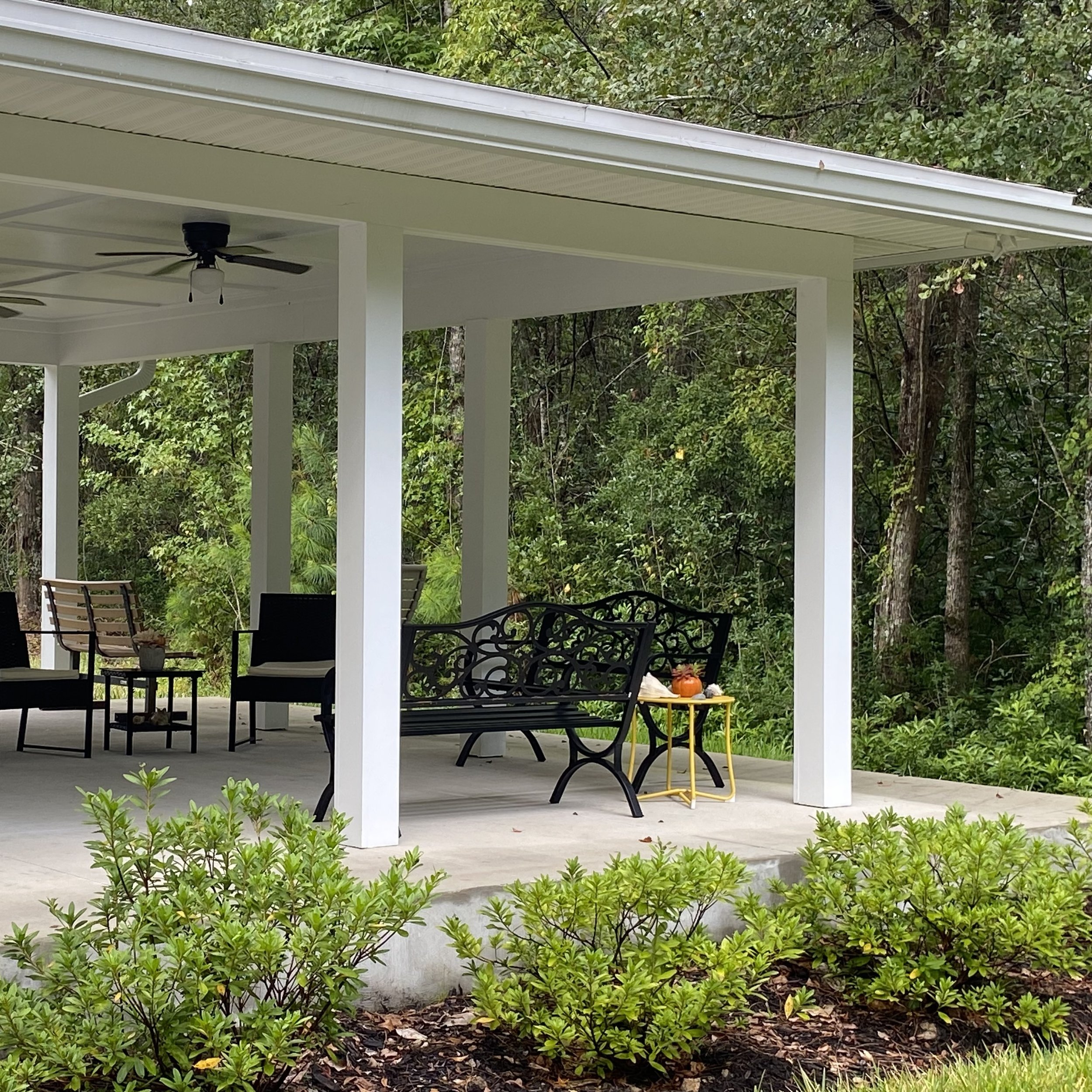 Outdoor pavilion with benches, small table, and ceiling fans, surrounded by trees and bushes.