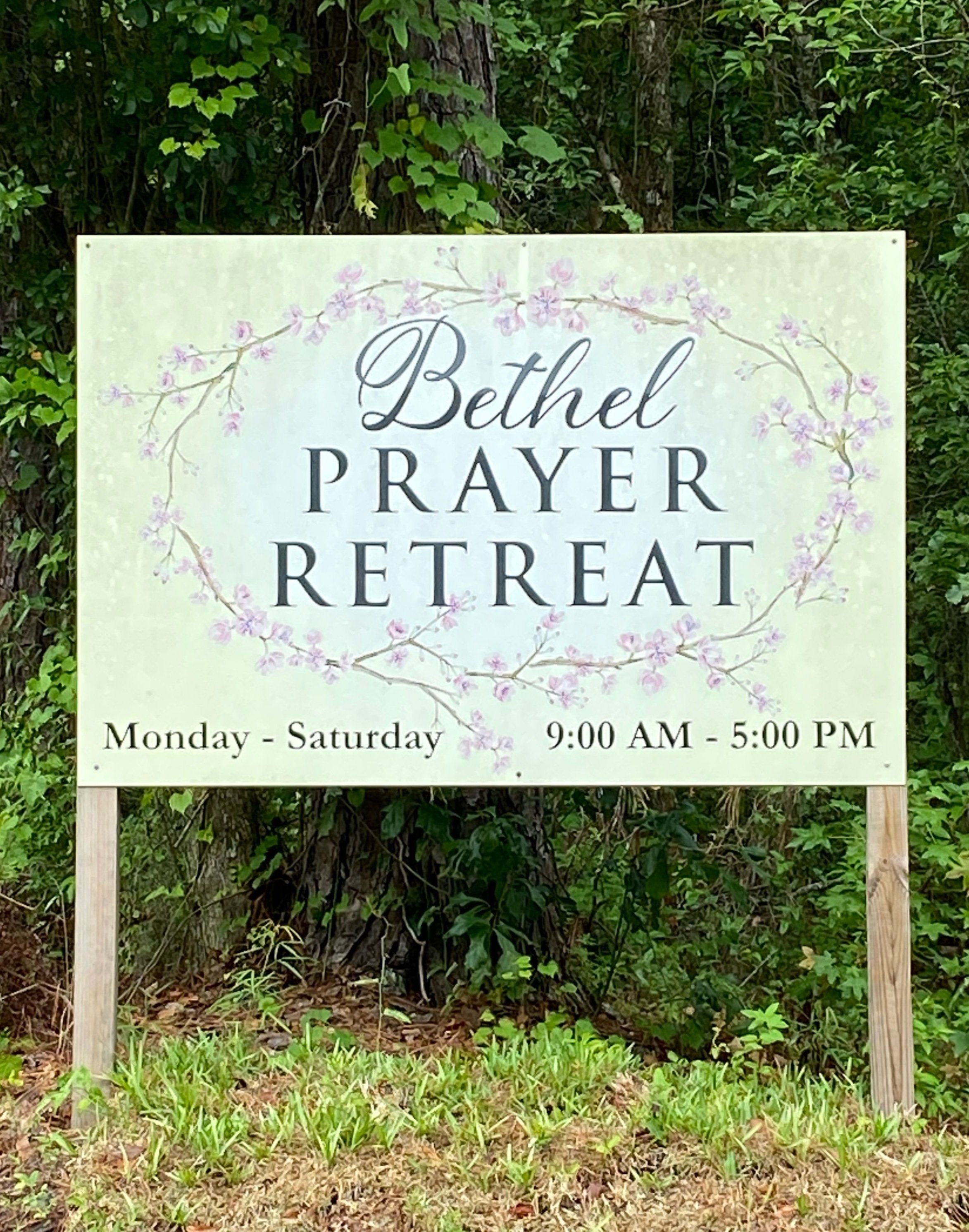 Outdoor sign for Bethel Prayer Retreat, open Monday to Saturday from 9:00 AM to 5:00 PM, surrounded by greenery and floral design.