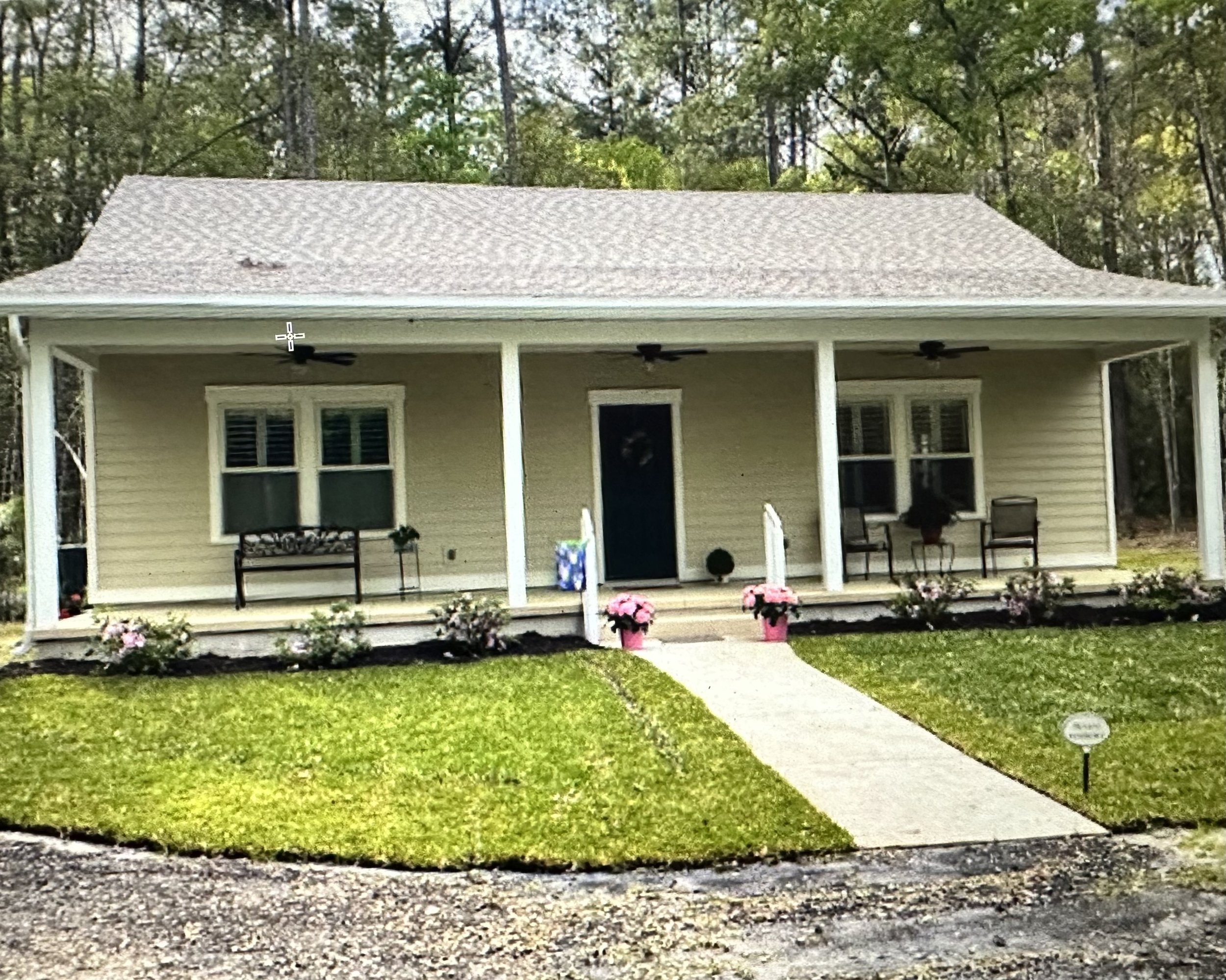 Small beige bungalow with front porch, two windows, and a well-maintained lawn with flowers and a pathway.