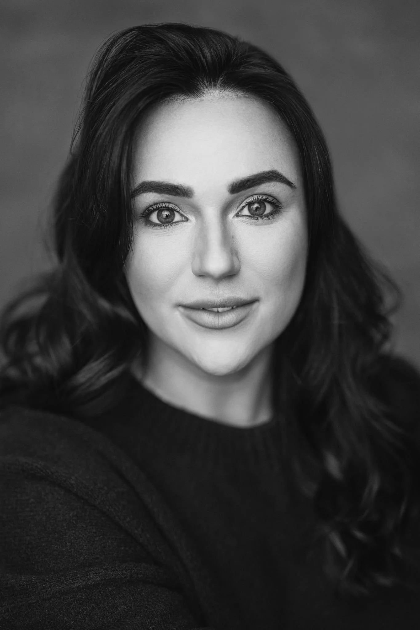 Close-up black-and-white portrait of a woman with long wavy hair, wearing a dark top, looking directly at the camera with a neutral expression.
