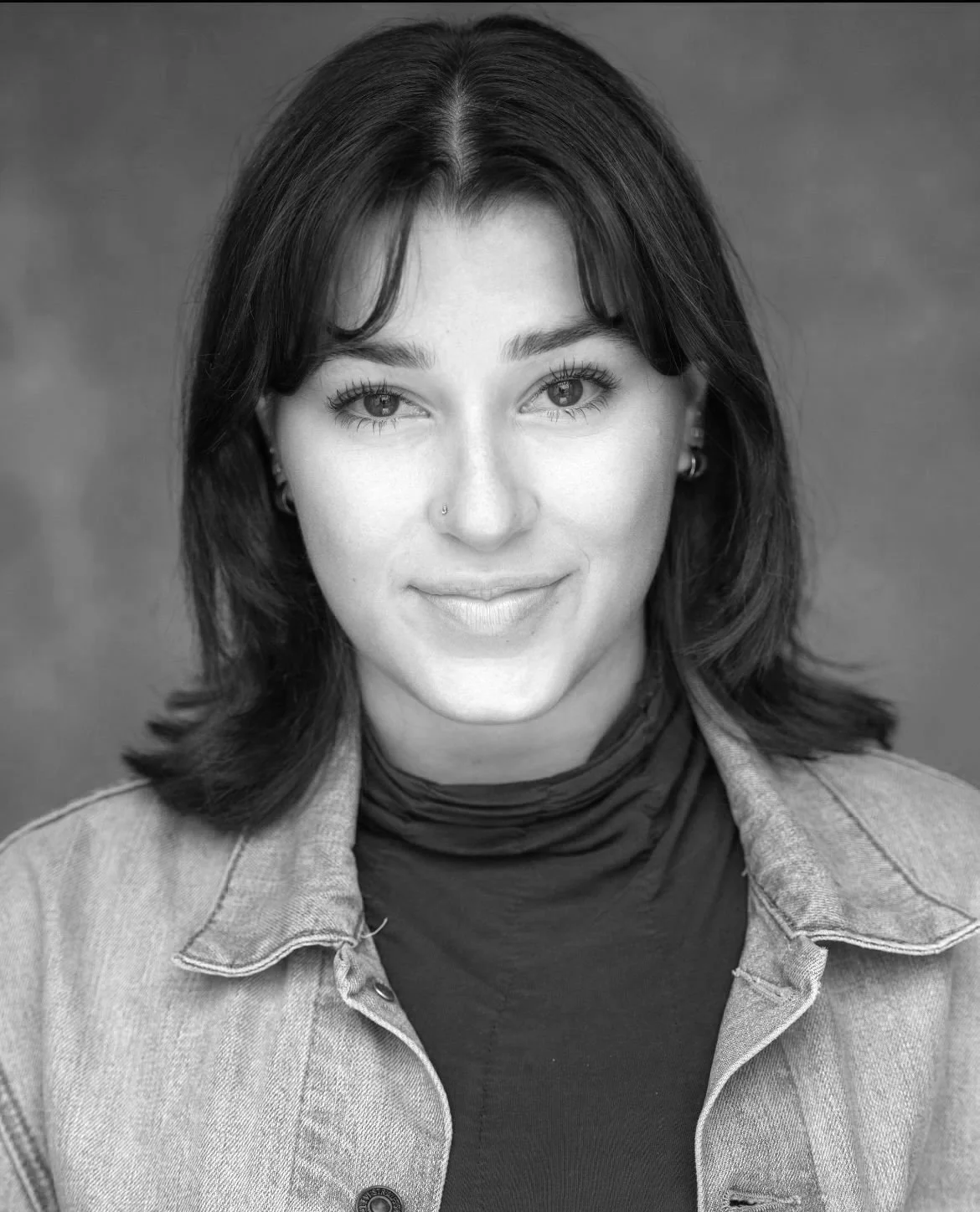 Black and white portrait of a young woman with shoulder-length hair, wearing a denim jacket over a turtleneck, smiling slightly.