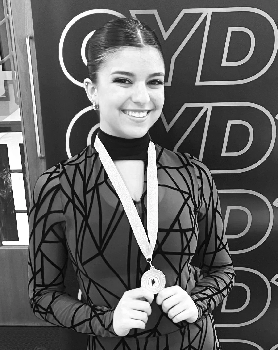 A woman smiling and holding a medal in front of a sign with the word 'CYD' in large letters.