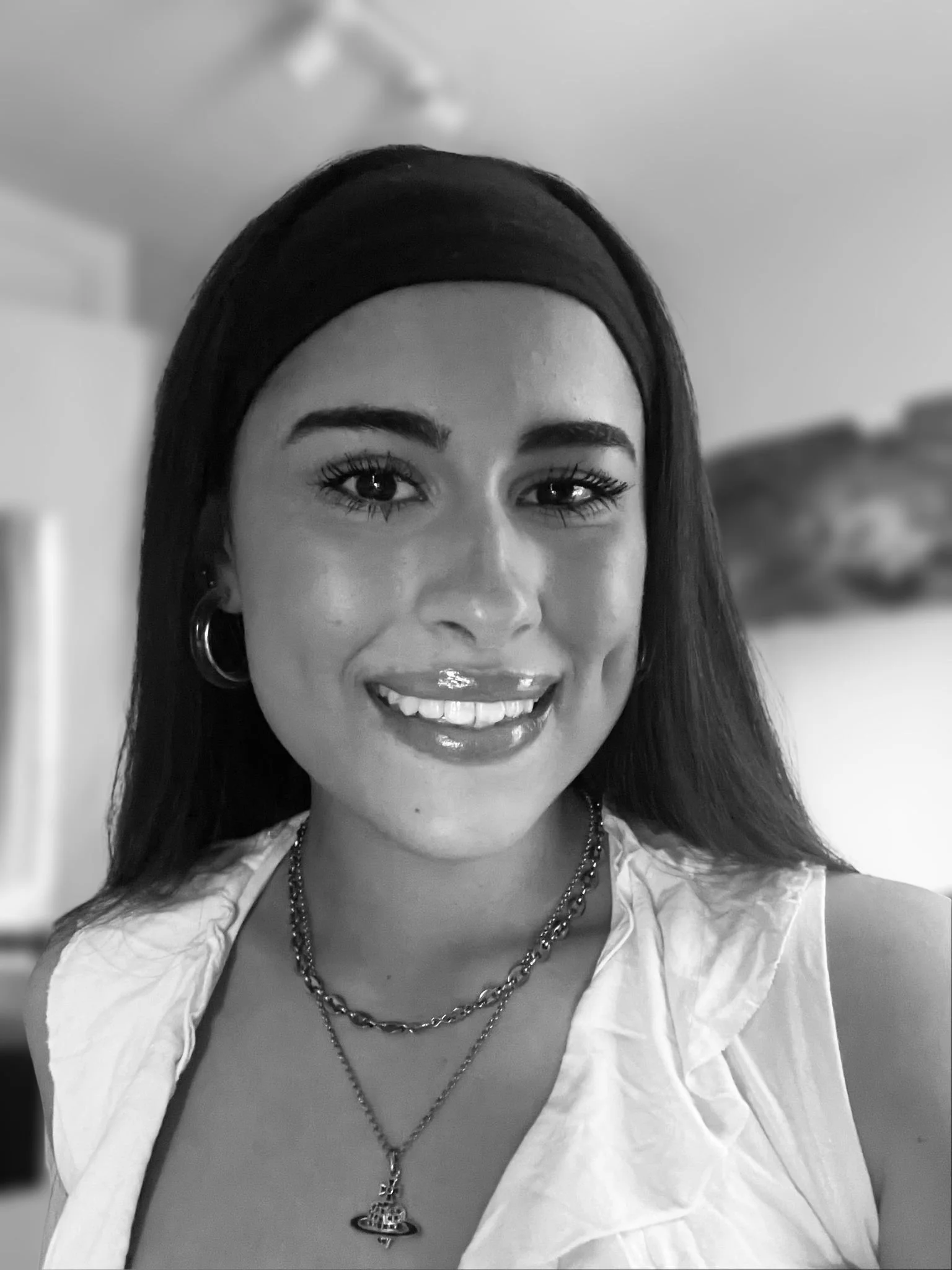A woman with long dark hair, wearing a headband, hoop earrings, layered necklaces, and a white sleeveless top, smiling in an indoor setting.