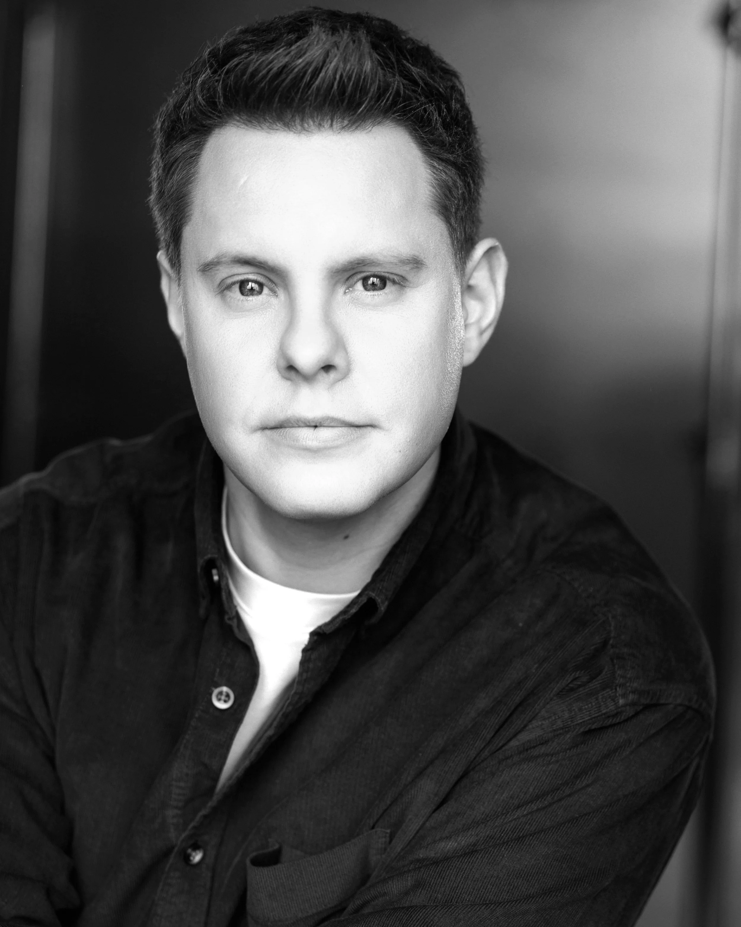 Black and white portrait of a young man with short hair, wearing a dark shirt and a white undershirt, looking directly at the camera with a neutral expression.