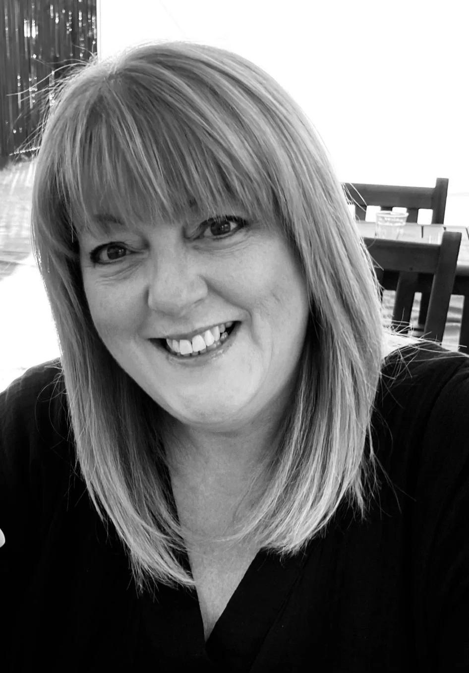 A smiling woman with straight, shoulder-length hair and bangs, wearing a dark top, sitting indoors with a dining area in the background.
