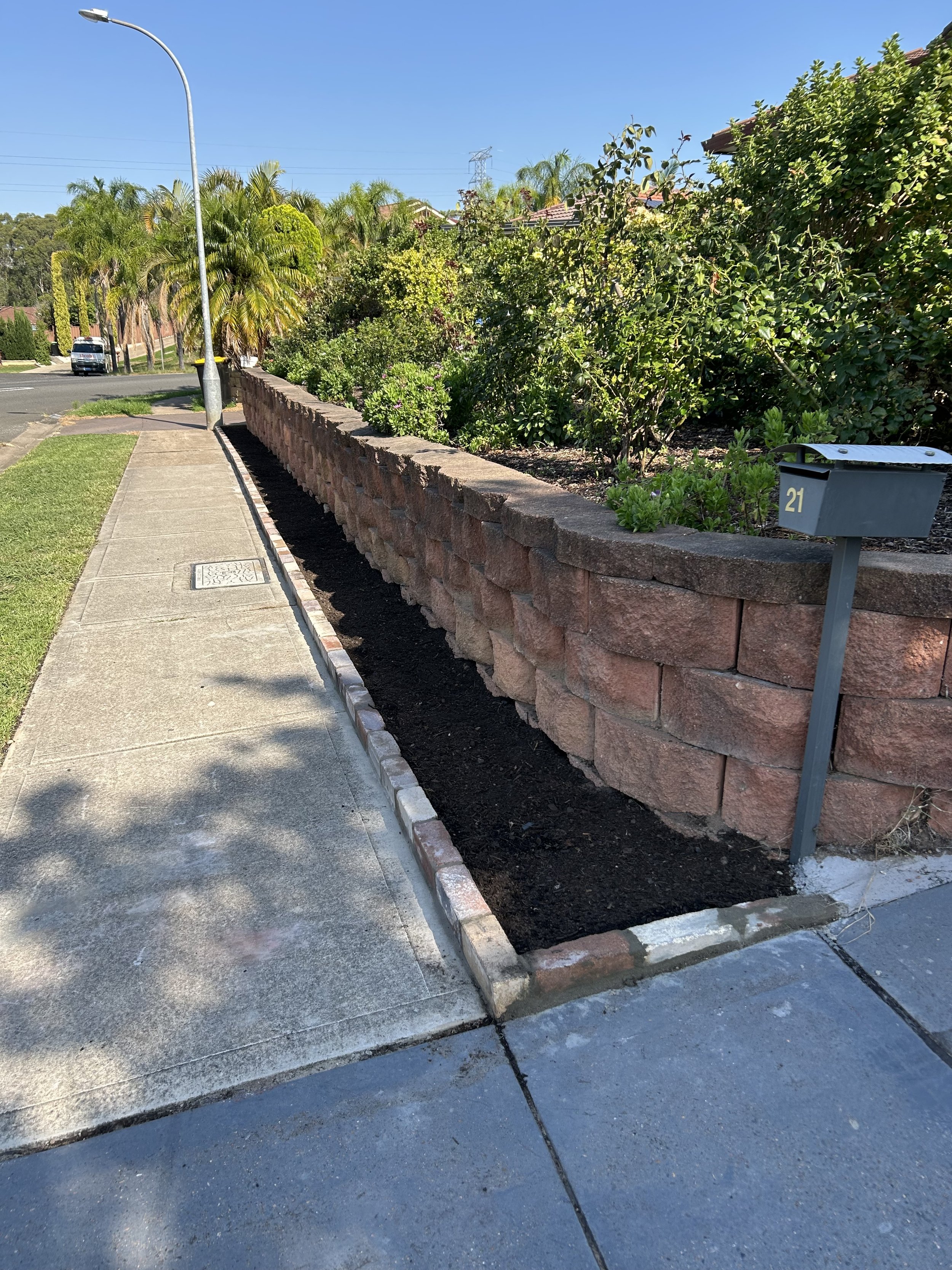 A sidewalk with a brick wall to the right, a newly planted garden bed with dark soil, a mailbox labeled '21' in front of the garden, a streetlamp, and lush green bushes and trees along the street under a clear blue sky.
