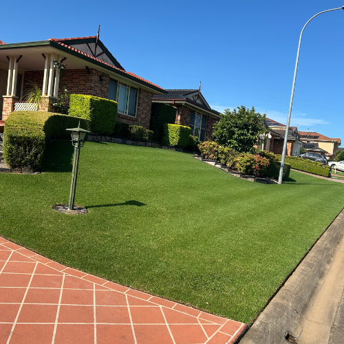 A suburban front yard with a well-maintained green lawn, a brick house with bushes and trees, a streetlamp, a paved sidewalk, and a clear blue sky.