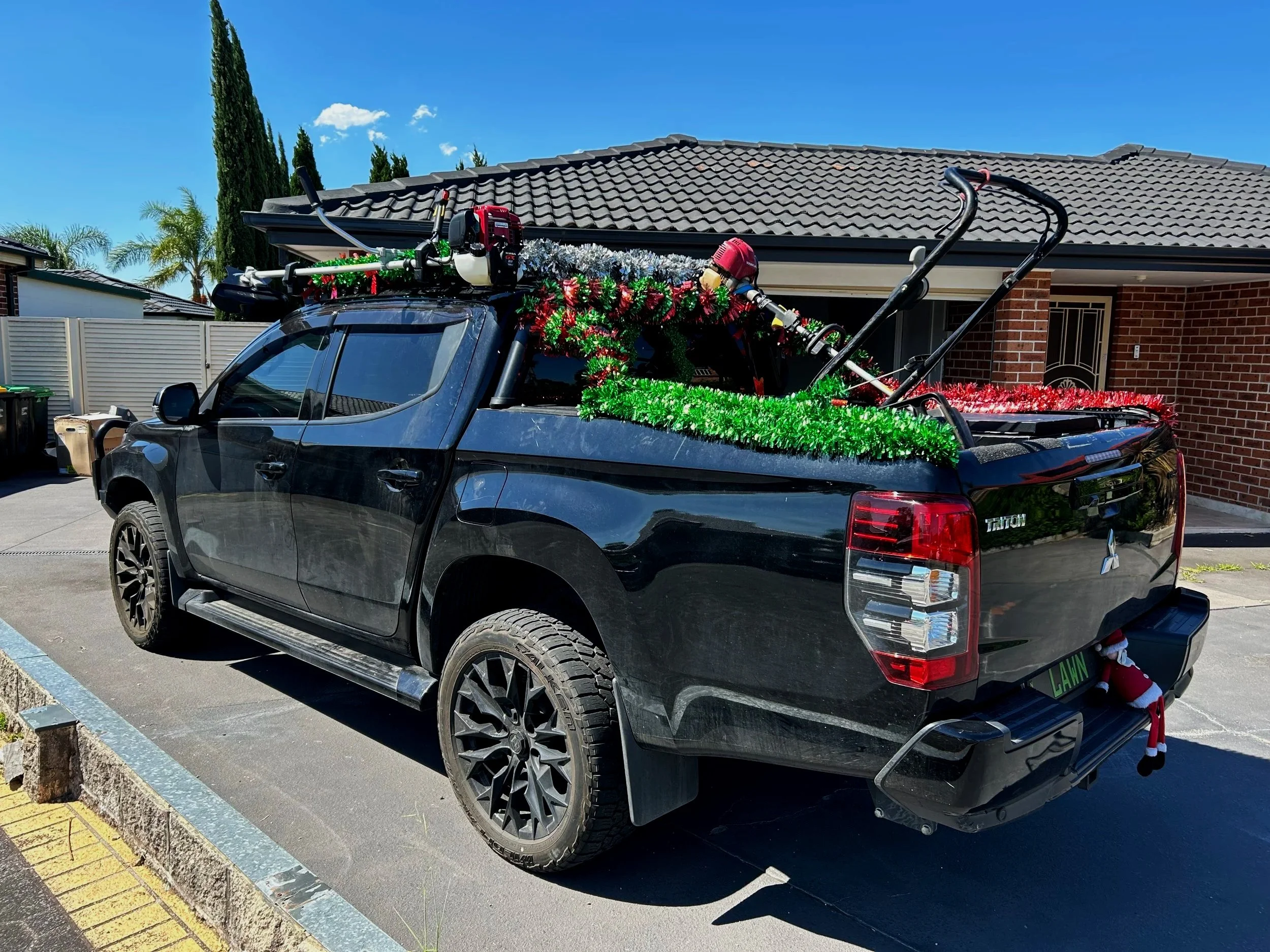 Black pickup truck decorated with Christmas tinsel and festive ornaments, parked in driveway in front of a brick house with a gray tiled roof and clear blue sky.