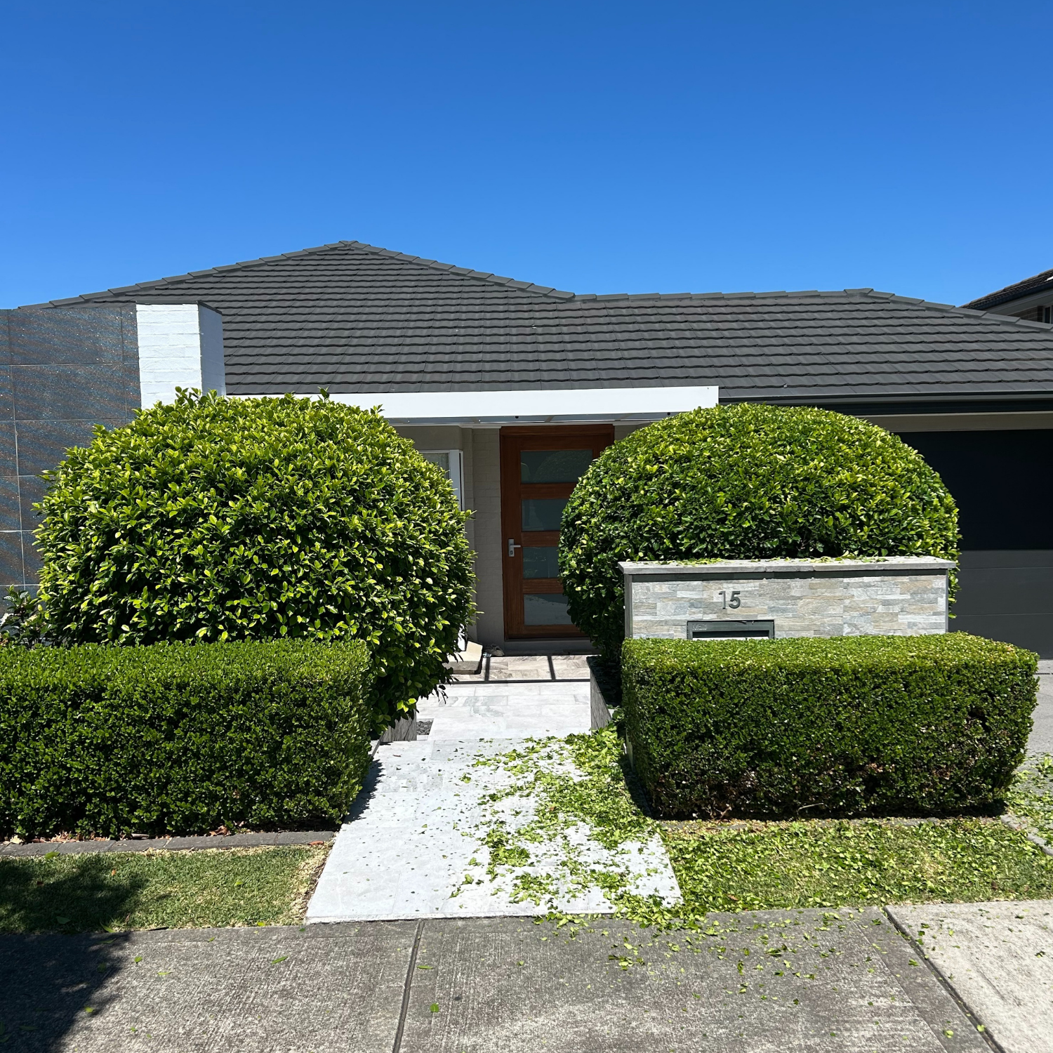 Front view of a house with a gray tiled roof, wooden front door, two large green bushes flanking the door, and a small hedge at the sidewalk. House number 15 displayed on a stone wall near the bushes, with a clear blue sky in the background.