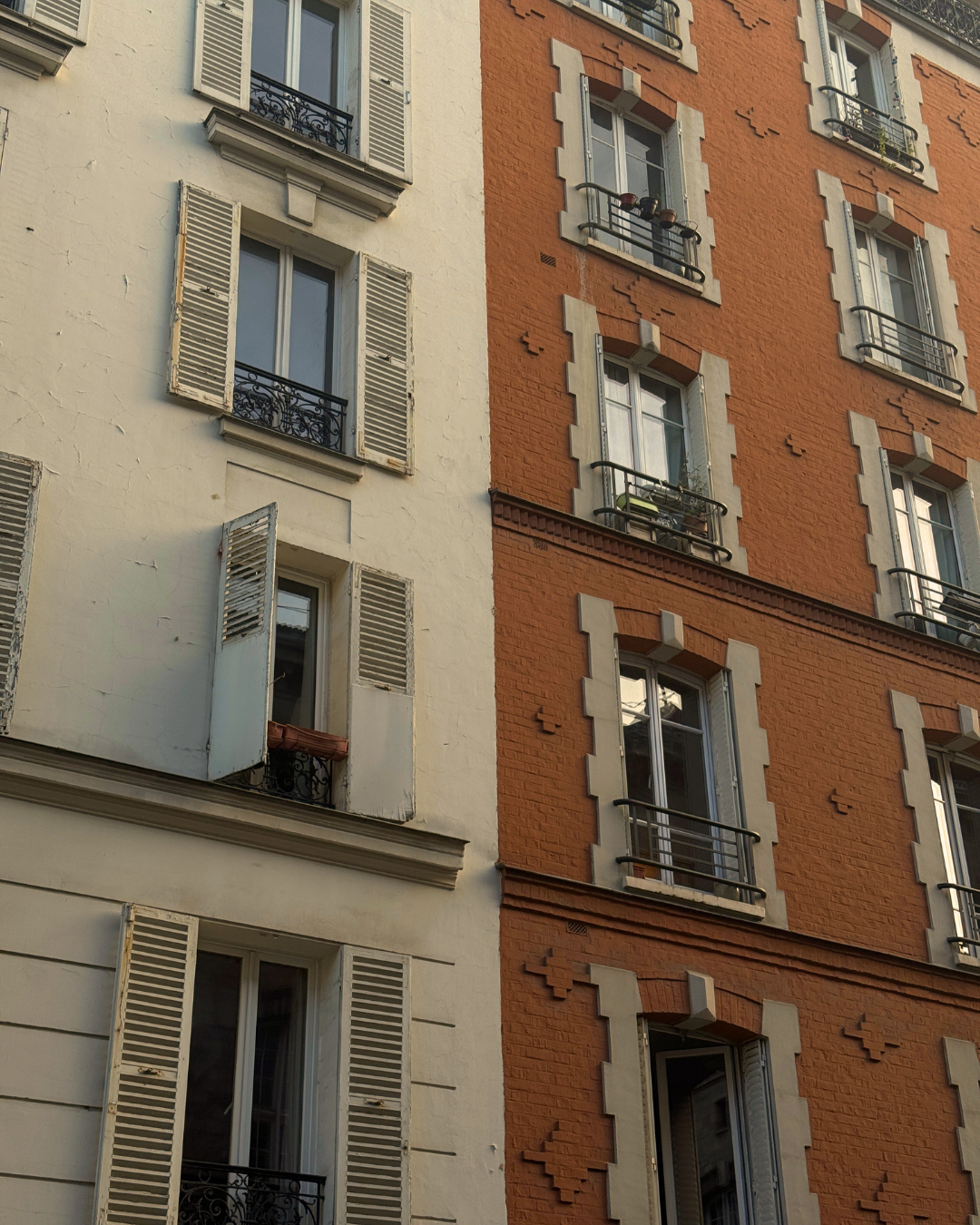 Close-up of the exterior of two adjacent buildings with European-style windows and shutters, one painted white and the other orange brick, with some open shutters and balcony railings.
