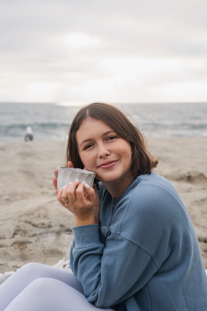 A woman sitting on a sandy beach, holding a ceramic bowl, with ocean and cloudy sky in the background.