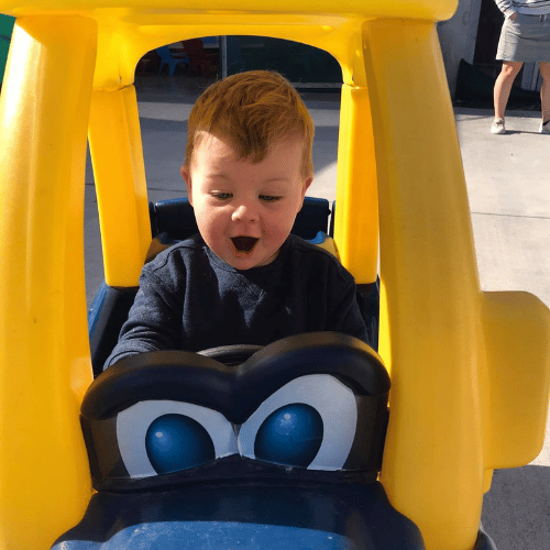 A young child beaming with joy while riding a yellow toy car outside at Happy Days Childcare, enjoying free play and active exploration.