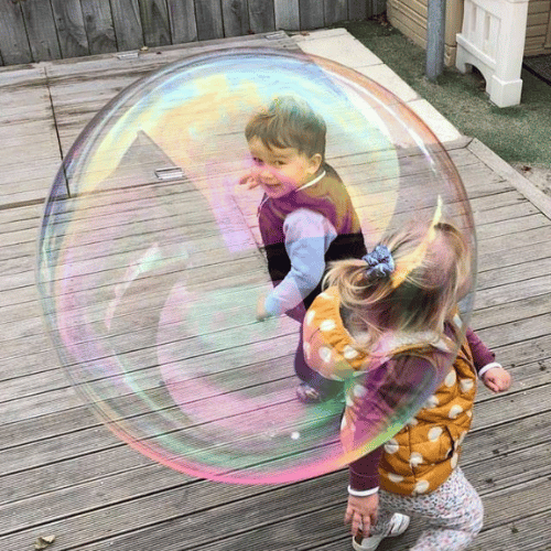Two young children laughing as they are surrounded by a giant bubble during a sensory and science play moment at Happy Days Taradale.