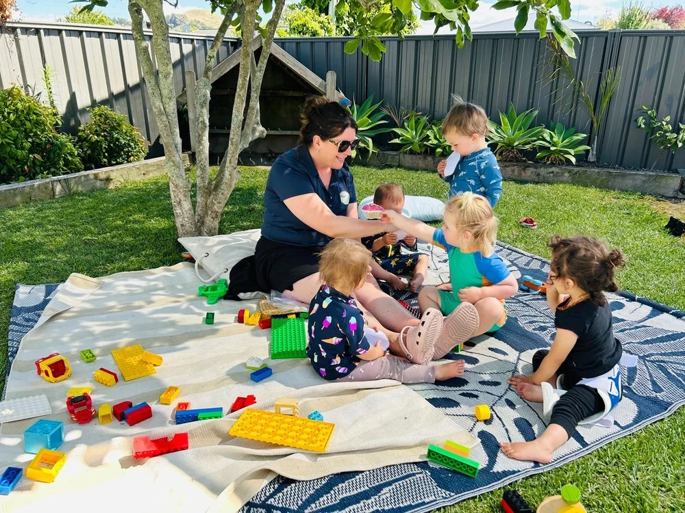 Teacher sitting on a blanket with toddlers playing with Lego and connecting in the shade at Happy Days Taradale