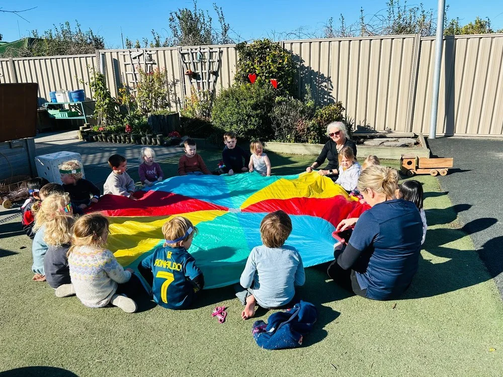 Children and teachers gathered around a colourful parachute for group play on the outdoor mat at Happy Days Childcare