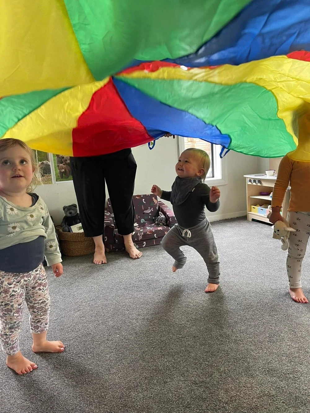 Excited toddler jumping under rainbow parachute with peers during indoor movement activity at Happy Days