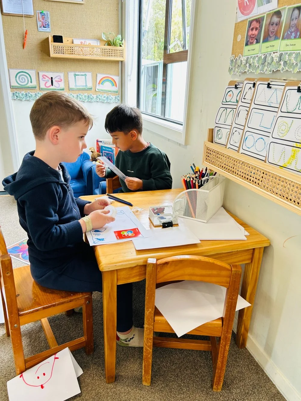 Two young boys working on literacy and drawing tasks at a classroom table in Happy Days' Puawai Room