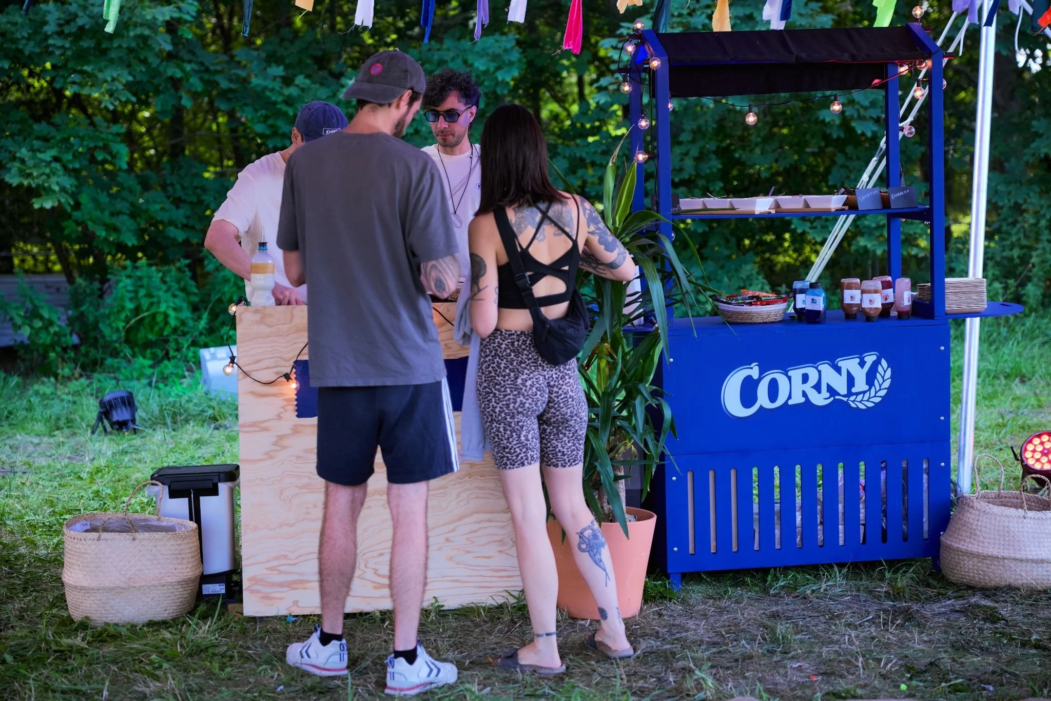 Menschen an einem Straßenstand mit der Aufschrift 'CORNY' bei einem Festival oder einer Veranstaltung im Freien, auf Grünflächen mit Bäumen im Hintergrund.
