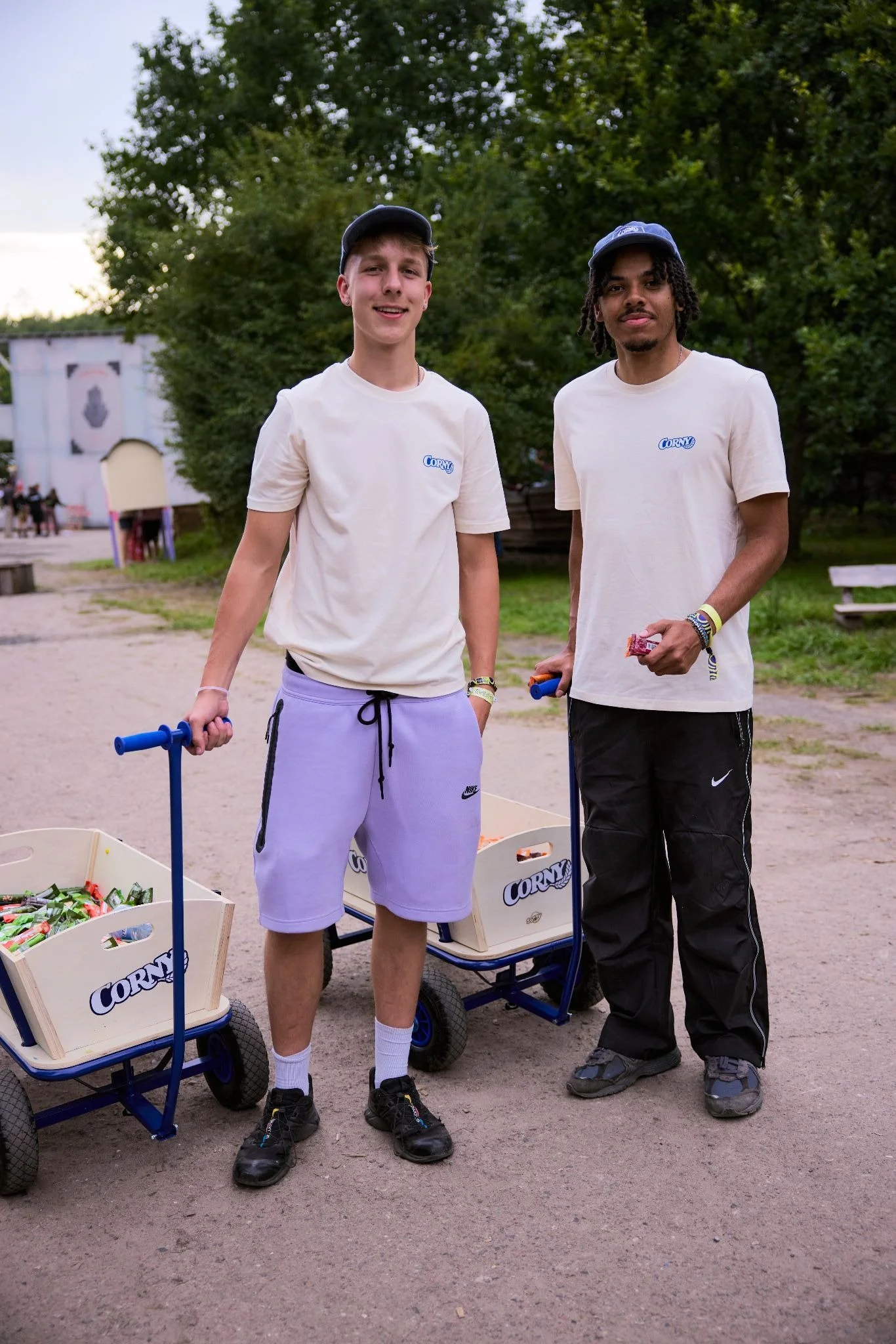 Zwei junge Männer stehen im Freien neben Wagen mit Snacks, tragen weiße T-Shirts mit 'CORN' Logo, und schauen in die Kamera.