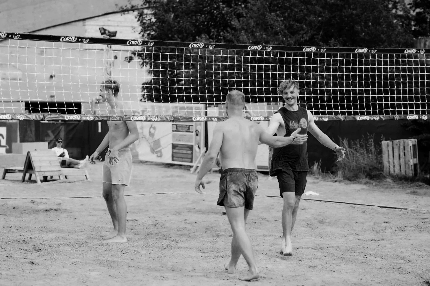 Drei junge Männer spielen Beachvolleyball auf einem Sandplatz. Einer gibt den Ball an einen anderen weiter, während ein dritter im Hintergrund sitzt.