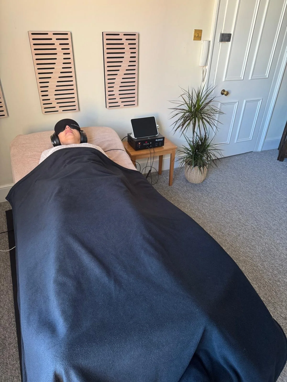 Person lying on a massage table with a black blindfold and headphones, covered with a black blanket in a room with beige walls, a small side table with electronic equipment, a potted plant, and a closed white door.