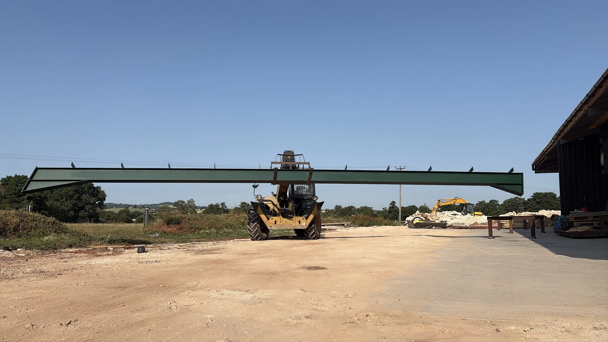 Construction site with a bulldozer lifting a green steel beam, a pile of rubble, and a partial building with a sloped roof on the right. Clear blue sky.