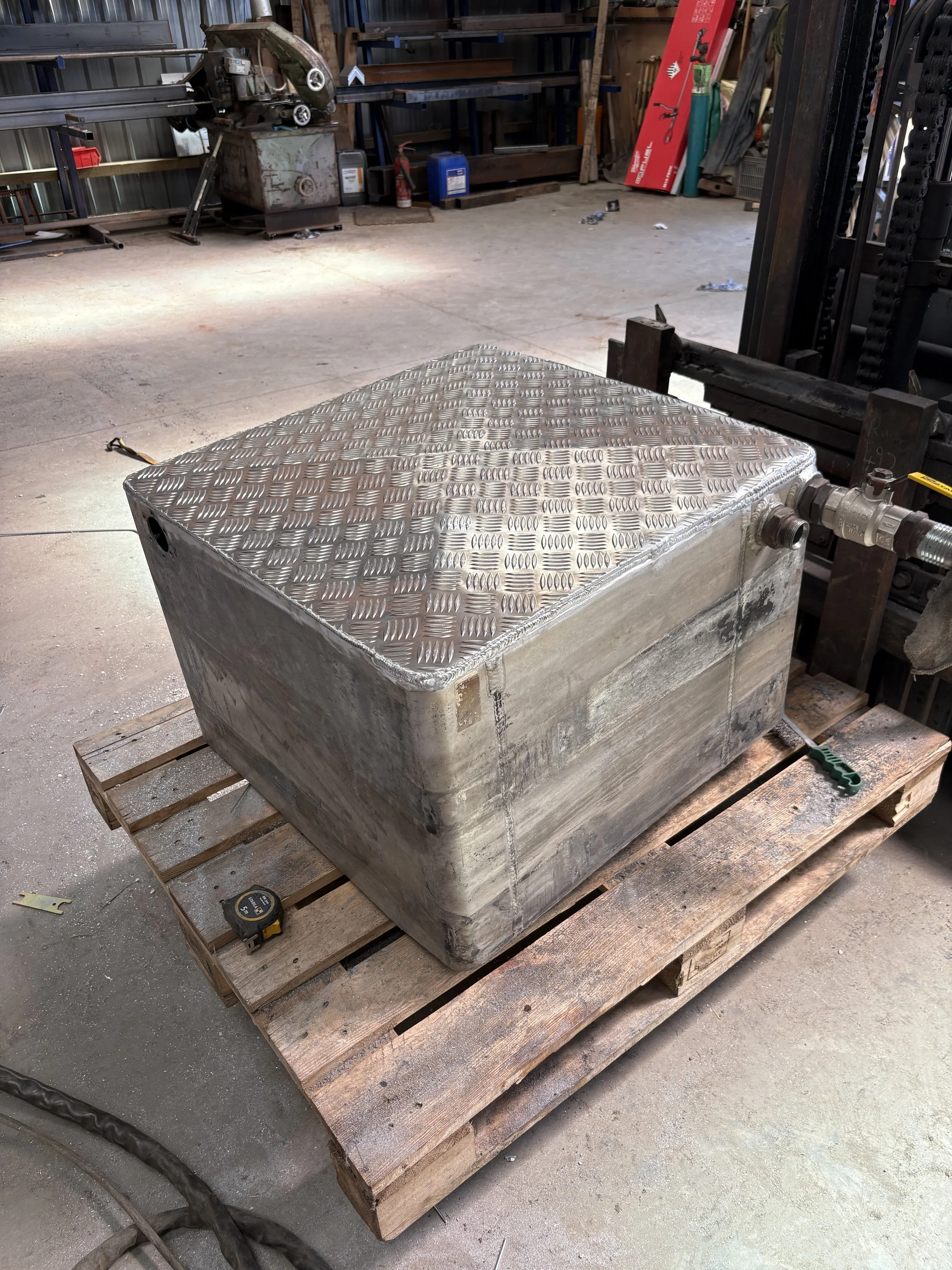 A large metal box with a textured, diamond plate top sits on a wooden pallet in an industrial warehouse or workshop. The surrounding area contains various tools, equipment, and storage shelves.