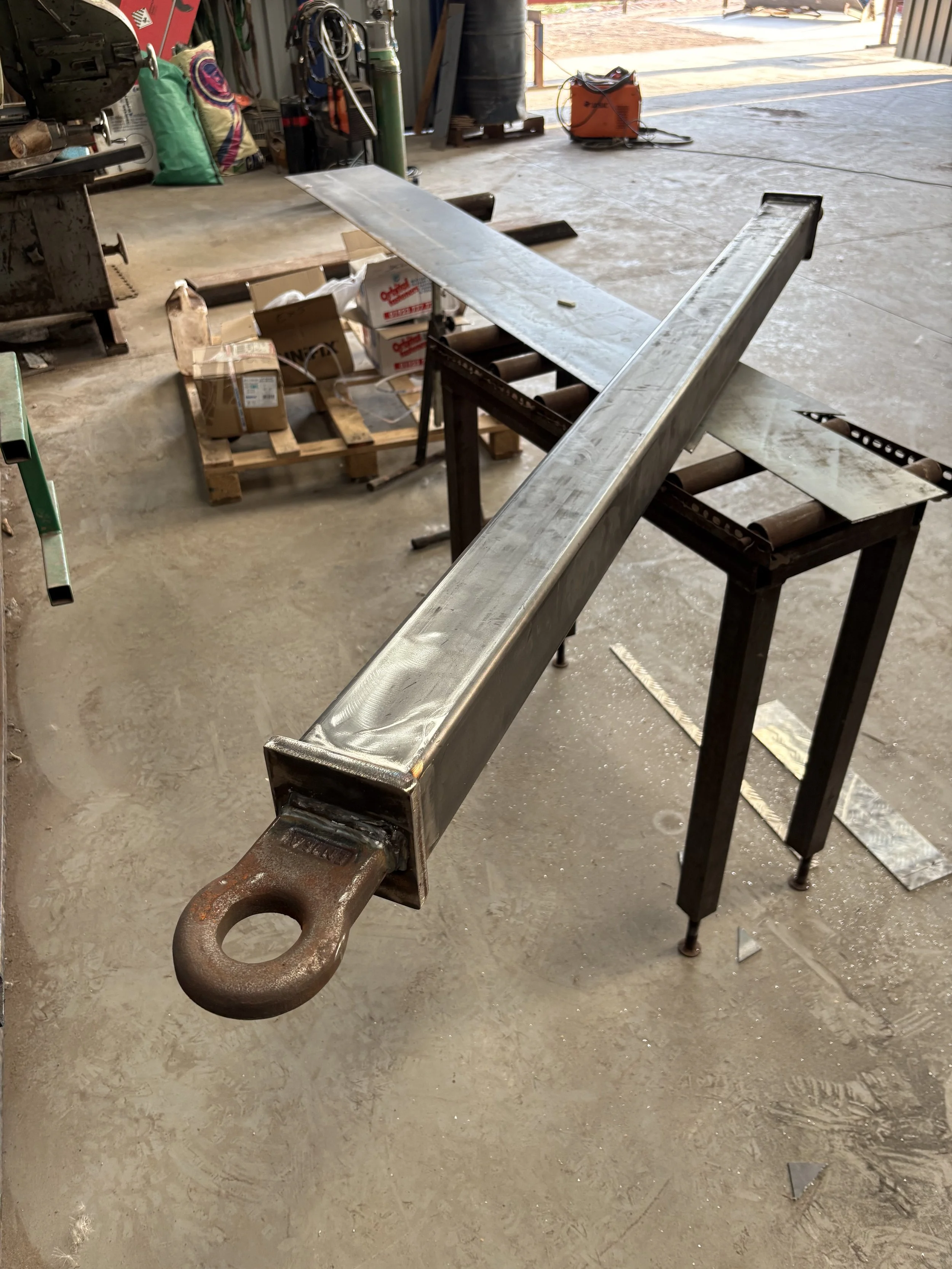 A metal crane hook attached to a CNC machine in a workshop or garage setting. The background includes a workbench with boxes and tools, and the floor appears to be unfinished concrete.