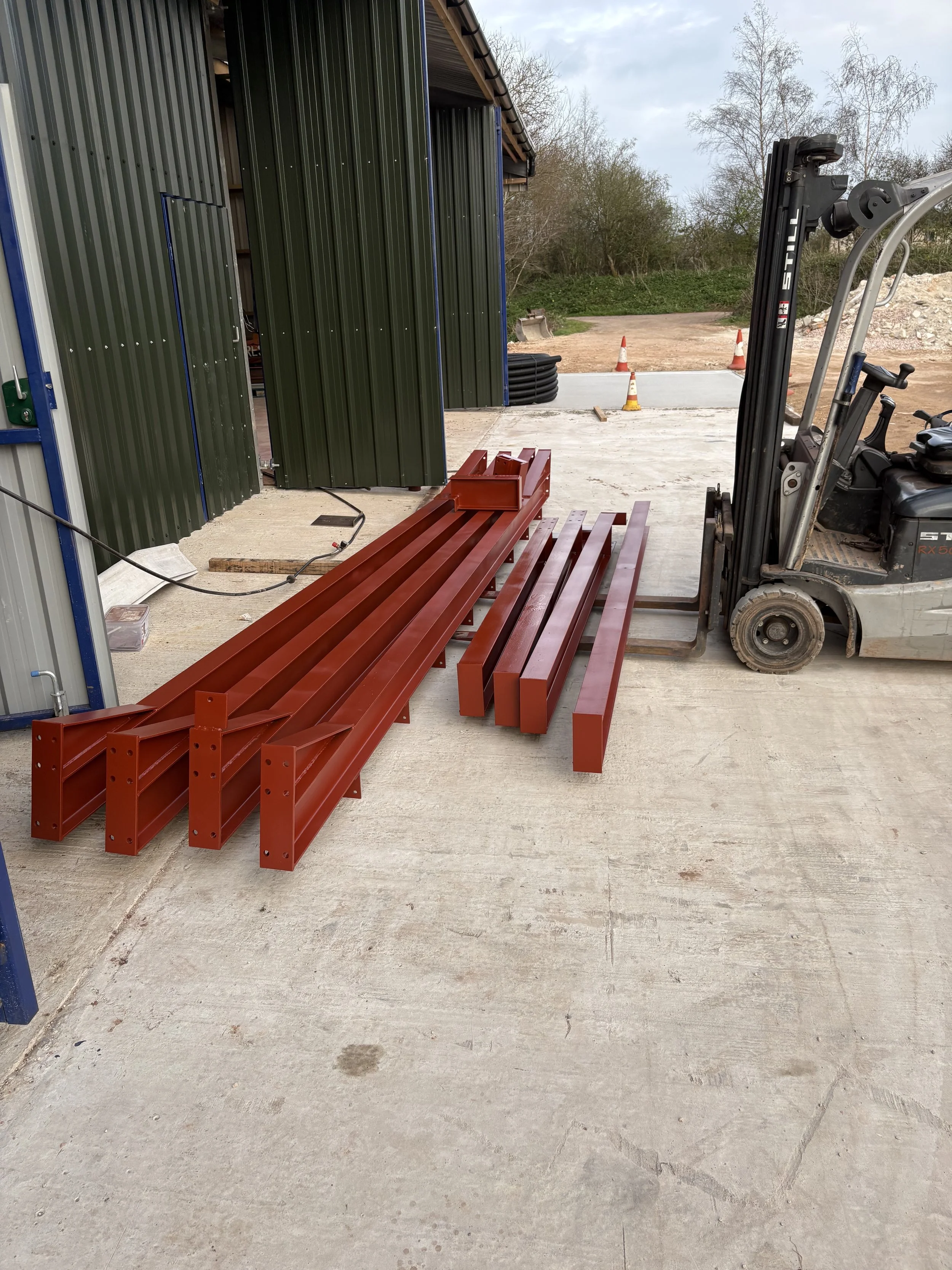 Construction site with red metal beams, a forklift, and traffic cones, next to a green storage shed and a concrete area with trees and a dirt road in the background.