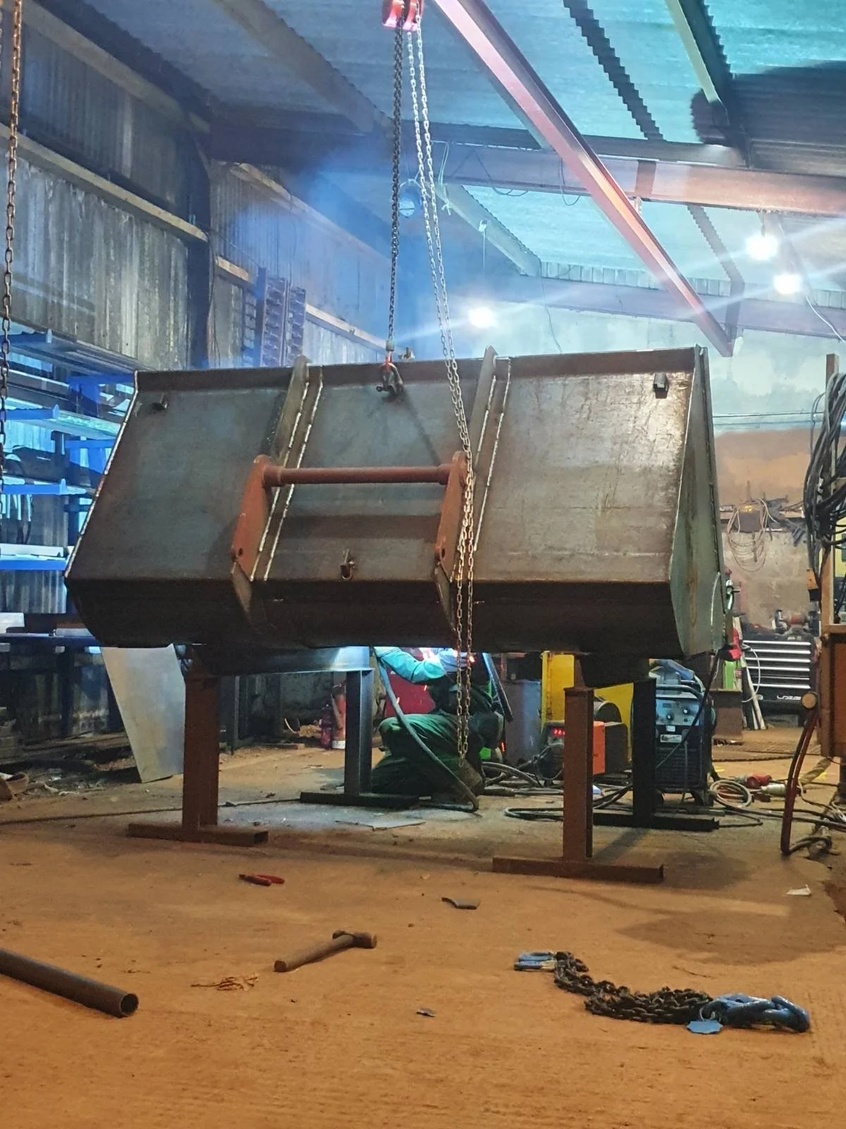 Large 2m3 telehandler bucket being welded inside an industrial workshop, suspended by chains, with various tools and equipment around.