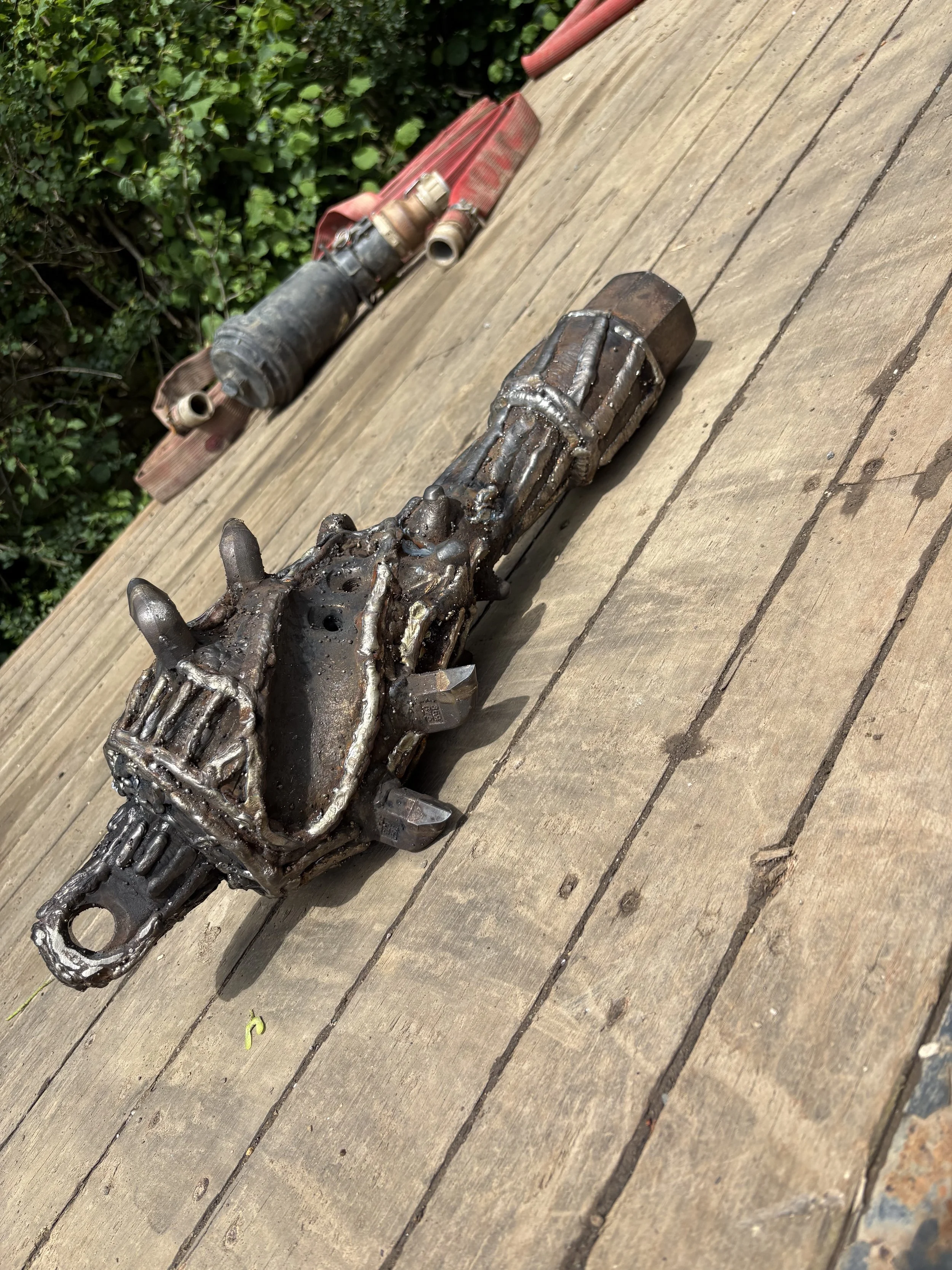 A metal mechanical object resembling a futuristic or steampunk-style gun or weapon, placed on a wooden deck near green bushes, with a red toolbox or container in the background.