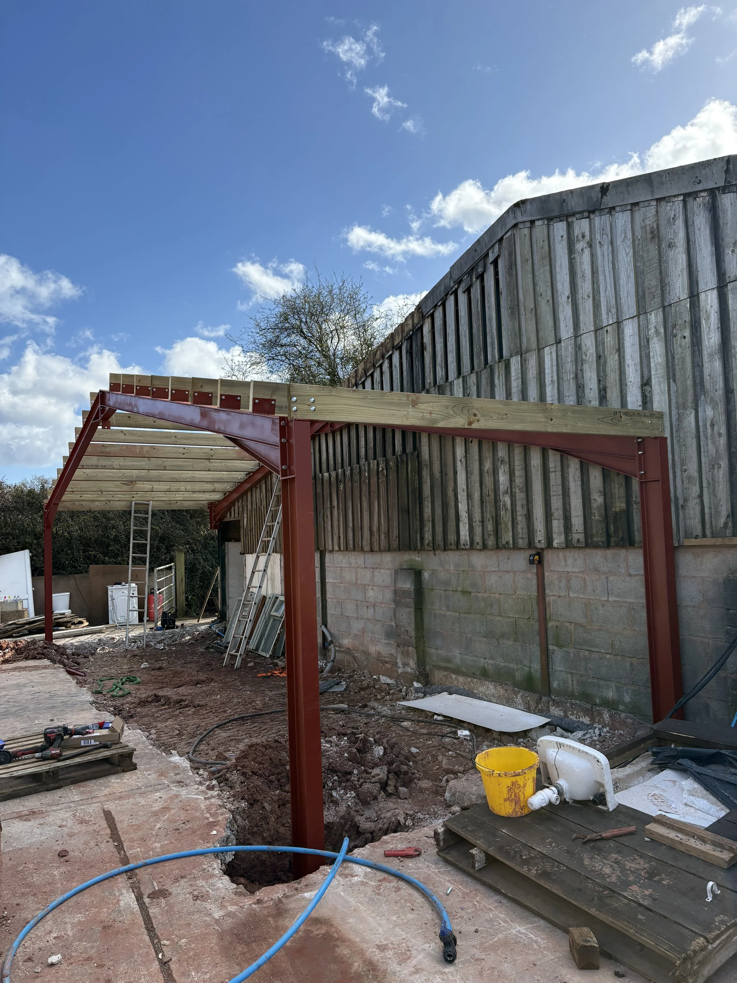 Construction site with partial steel framework, ladders, and tools, next to a wooden fence and brick wall under a blue sky with clouds.