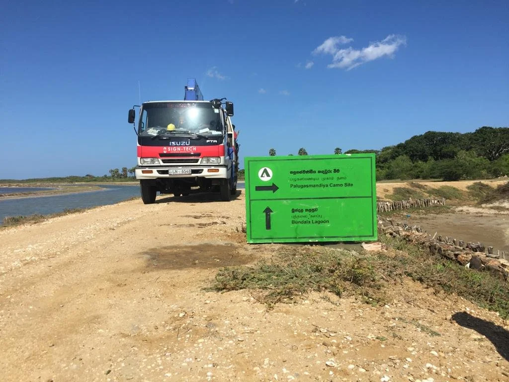 A dirt road near a body of water with a truck and a green directional sign indicating Palugasmandiya Camp Site to the right and Bundala Lagoon straight ahead.