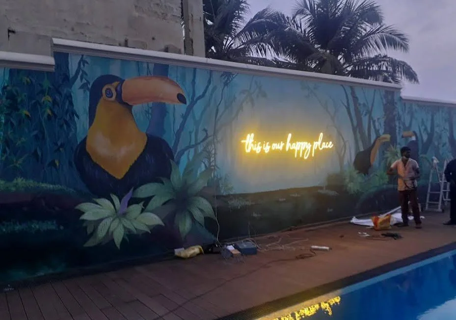 Colorful mural of a toucan on a jungle background with a neon sign that reads 'this is our happy place' next to a pool, with two people working on the mural.