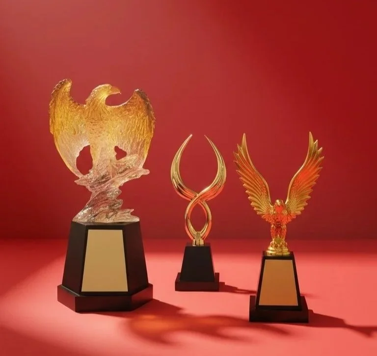 Three ornate trophies with gold and black bases are displayed on a red background. The left trophy features a golden eagle with outstretched wings, the middle one has a sleek gold and transparent design with curved elements, and the right trophy depicts a golden eagle with detailed feathers and wings spread upward.