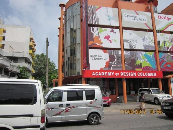 Exterior of the Academy of Design Colombo building with a large mural on the front, cars parked in front, and people walking nearby.