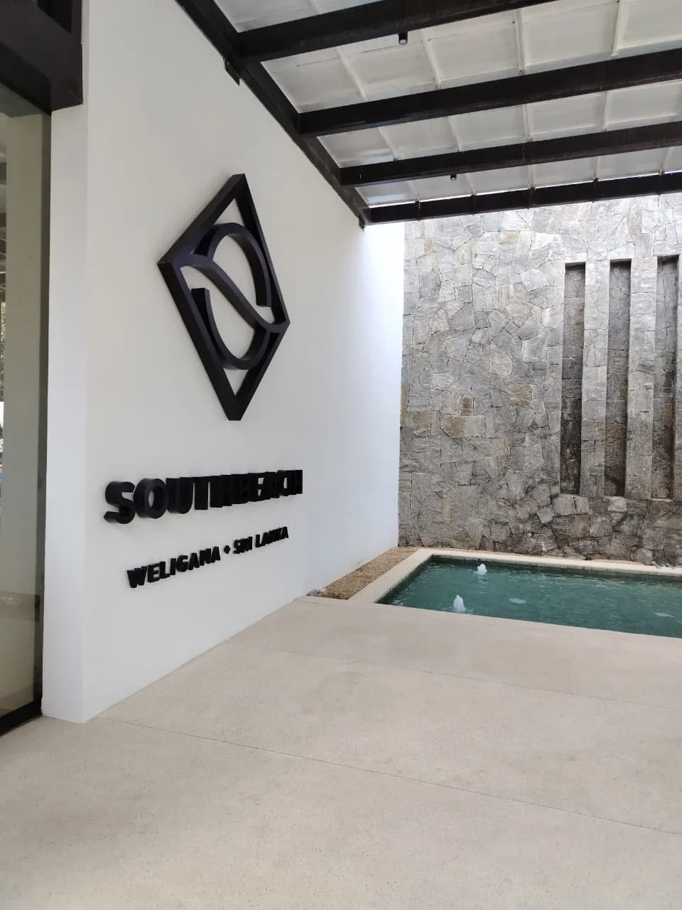 Outdoor area with a white wall displaying black 3D sign and text, a water feature, and a stone wall backdrop with vertical niches.
