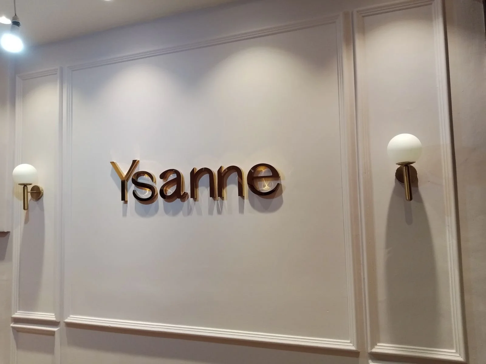 Interior wall with the word 'Y-sanne' in large, gold-colored letters, flanked by two wall-mounted light fixtures with white globes.