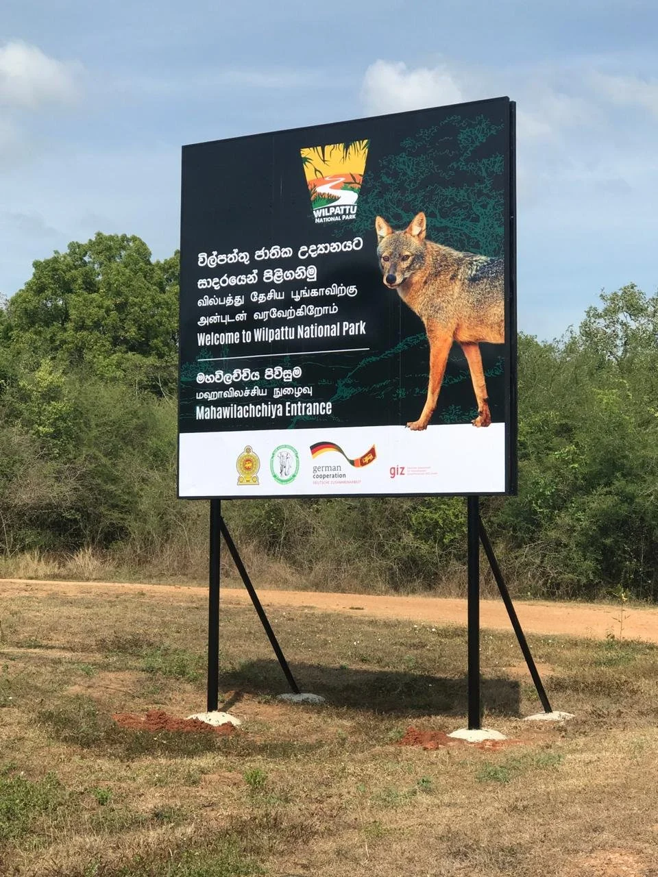 Signboard welcoming visitors to Wilpattu National Park with text in multiple languages, a logo of the park, logos of supporting organizations, and an image of a wild dog.