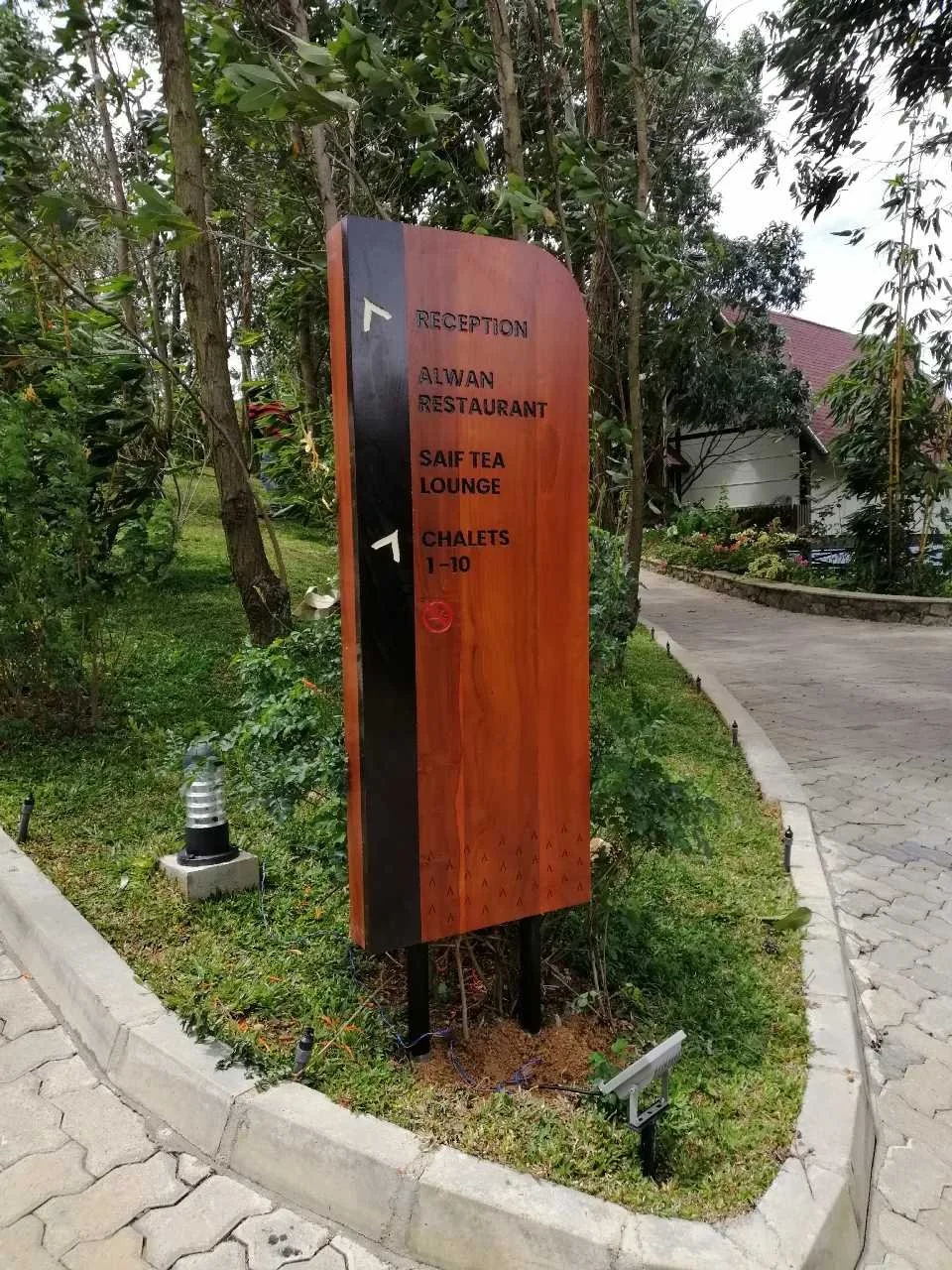Wooden directional signpost with lettering pointing to reception, Aiwon restaurant, Saif Tea Lounge, and chalets 1-10, located on a pathway surrounded by trees and greenery.