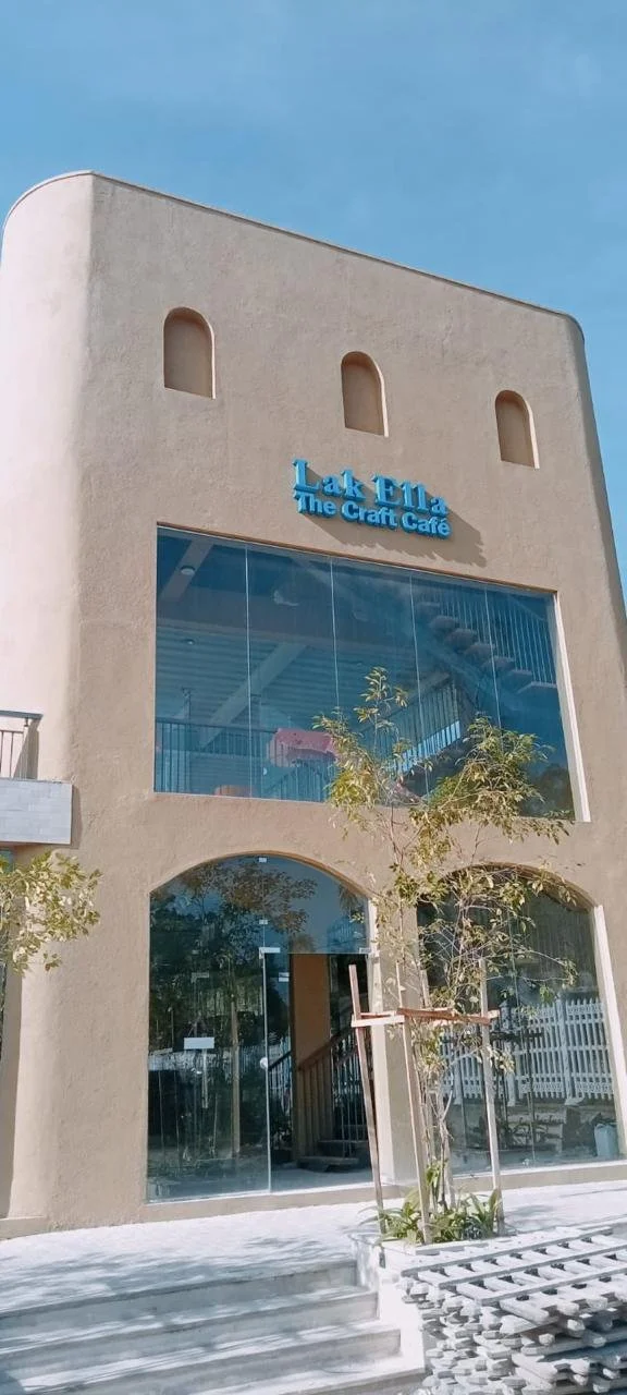 A modern beige building with a large glass window on the second floor, a small tree in front, and a sign that reads 'Lake Elma The Craft Café' in blue letters.