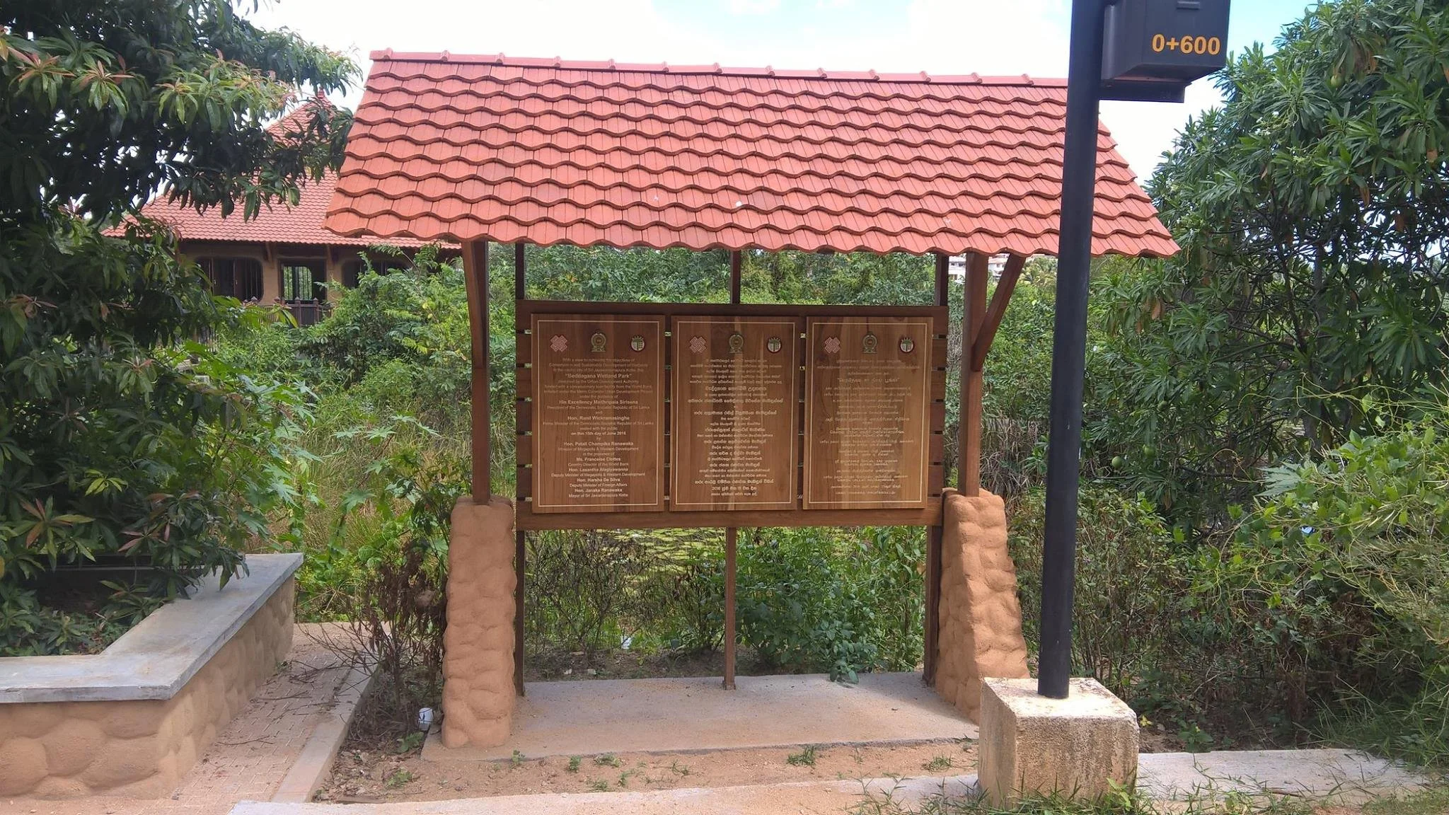 A wooden informational signboard with a red tiled roof, situated outdoors surrounded by greenery, with a pathway and a black post nearby.