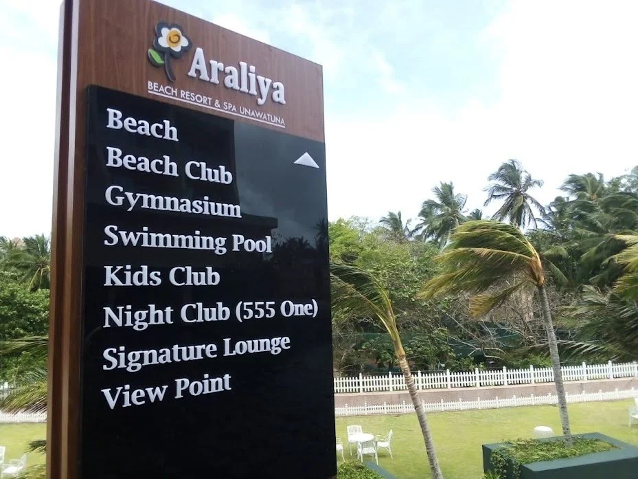 Sign for Araliya Beach Resort & Spa Unawatuna listing amenities like beach, beach club, gymnasium, swimming pool, kids club, night club, signature lounge, and view point, with tropical palm trees and outdoor seating in the background.