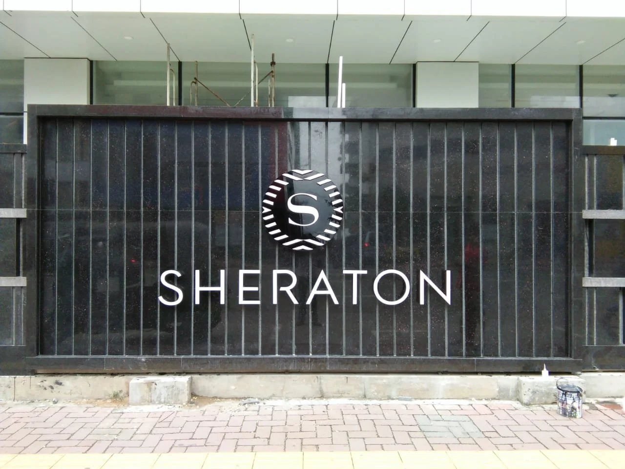Hotel sign displaying the Sheraton logo and name on a black metal fence, with construction scaffolding and building windows in the background.