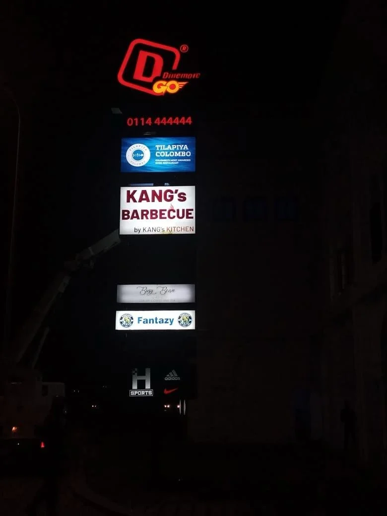 Nighttime photo of a vertical advertising signboard with multiple illuminated business signs including a restaurant, a football academy, a barbecue place, a cafe, a sports shop, and popular brand logos.