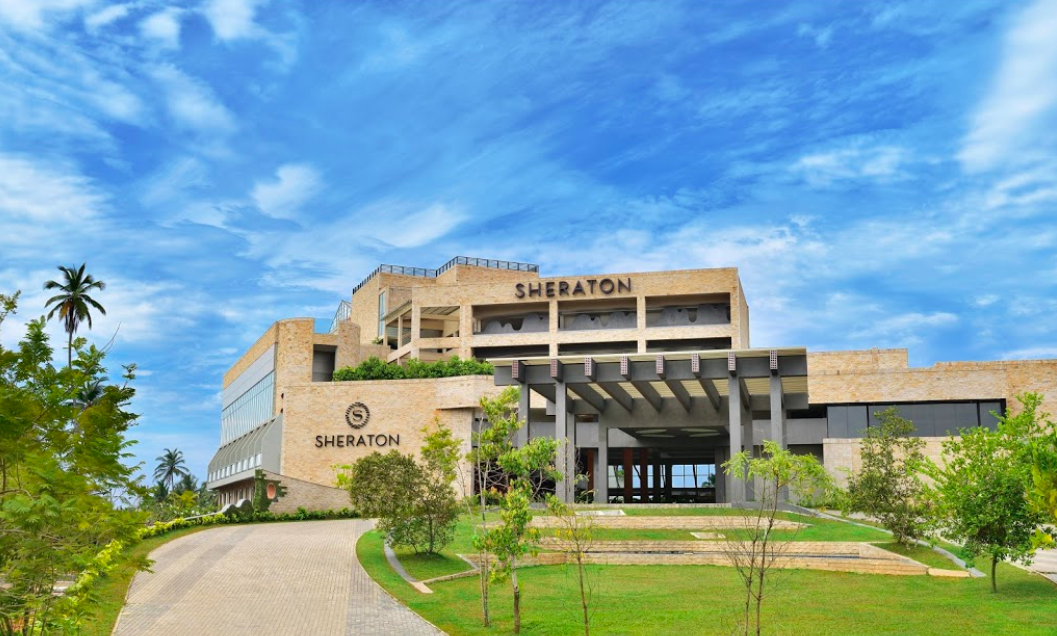 Exterior view of a Sheraton hotel building surrounded by green trees under a partly cloudy blue sky.